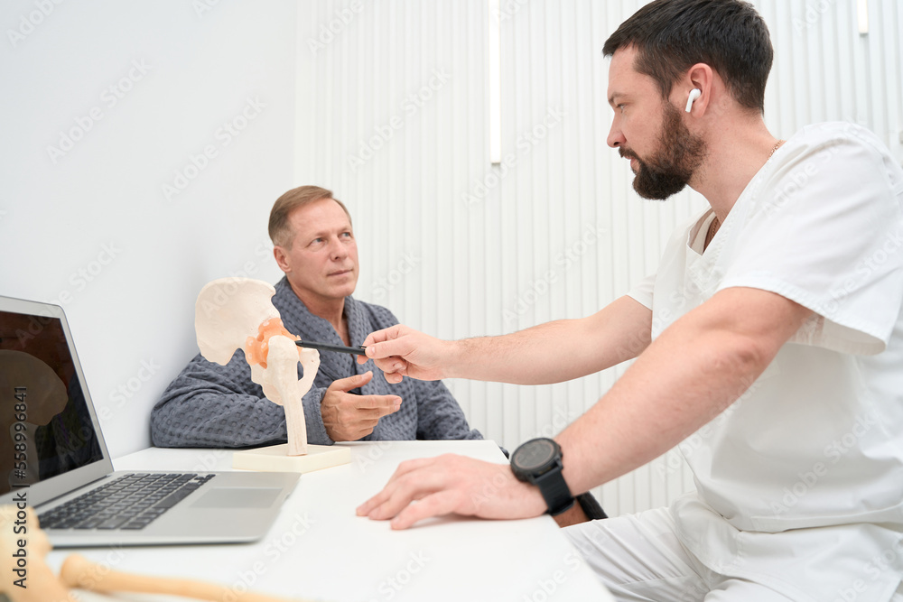 Certified doctor explaining hip-bone anatomy to middle-aged man Certified doctor explaining hip-bone anatomy to middle-aged man