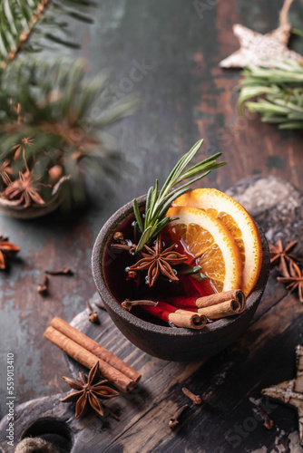 Mulled red wine in ceramic bowl close up with spices, fruits and rosemary on wooden rustic table, dark key. Traditional hot drink at Christmas time and winter