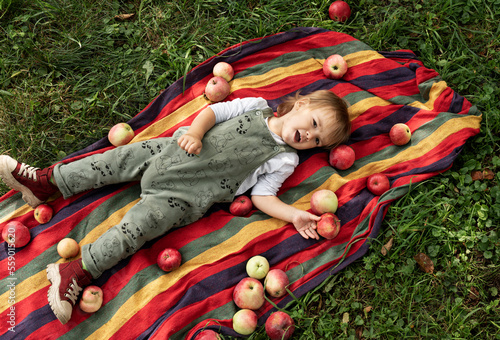 Slika na platnu A small child lies in the garden on a bedspread, apples are scattered around