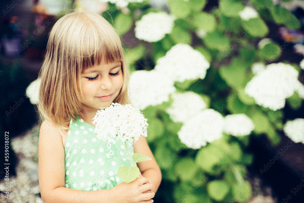A little girl in a beautiful dress with flowers and short blonde hair enjoys beautiful flowers in her grandmother's garden in the village A little girl in a beautiful dress with flowers and short blonde hair enjoys beautiful flowers in her grandmother's garden in the village