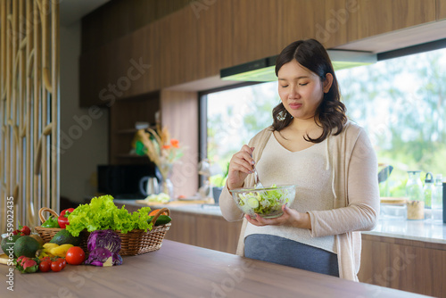 Beautiful Asian Pregnant Woman Happily Preparing a Vegetable Salad, Organic Healthy Food, in a Cozy Home Kitchen.