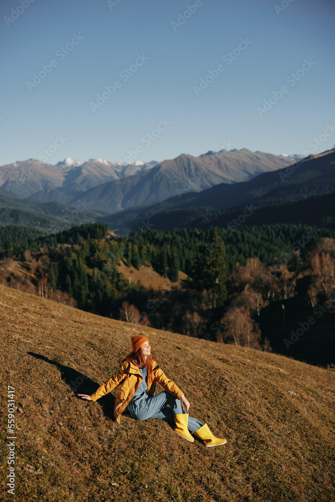 Woman full-length sitting resting on a hill smiling with teeth and looking at the mountains in a yellow raincoat and jeans happy hiking trip in winter, freedom lifestyle 