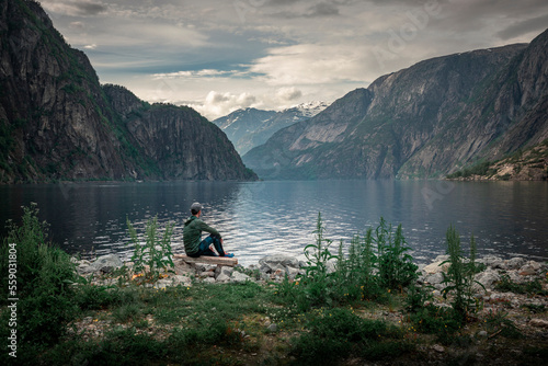 Man sitting at waterfront of lake in the mountain landscape Eidfjord in Norway  looking into the fjord  clouds in the sky