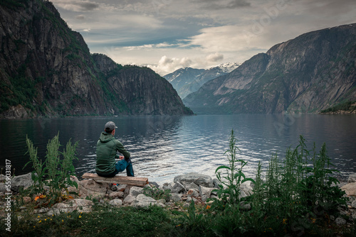 Man sitting at waterfront of lake in the mountain landscape Eidfjord in Norway, looking into the fjord, clouds in the sky