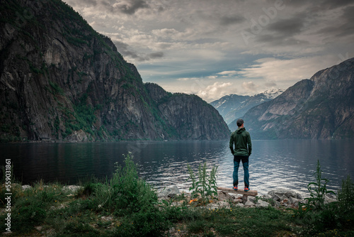 Man standing at waterfront of lake in the mountain landscape Eidfjord in Norway, looking into the fjord, clouds in the sky