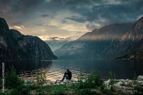 Man sitting at waterfront of lake in the mountain landscape Eidfjord in Norway, looking into the fjord, clouds in the sky during sunset with sun beams
