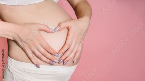 A young adult pregnant woman in white clothes touches her big naked belly with her hands. Makes a heart with his fingers. The concept of pregnancy. Close-up. Isolated on a pink background. A place for