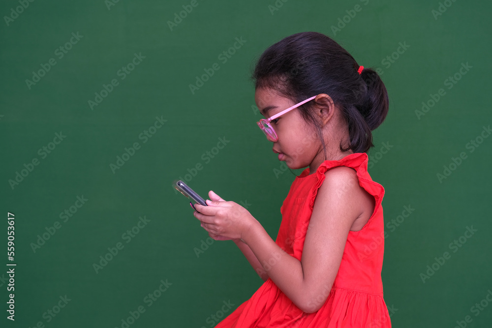 Little girl in her red dress and glasses busy playing game on her electronic gadget. Photo shot over dark green background.