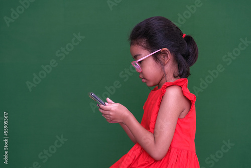 Little girl in her red dress and glasses busy playing game on her electronic gadget. Photo shot over dark green background. photo
