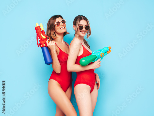 Two beautiful sexy smiling hipster women in red summer swimwear bathing suits. Trendy models having fun in studio. Hot female on blue. Cheerful and happy.  Playing toy water gun