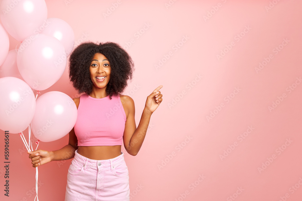 Pretty young and positive lady, stands isolated with bunch of pink balloons, pointing a side and smiles cheerfully Pretty young and positive lady, stands isolated with bunch of pink balloons, pointing a side and smiles cheerfully