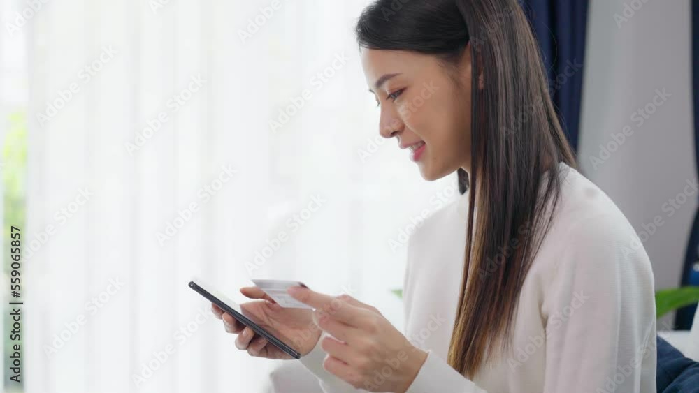 Young asian woman sitting on sofa in living room, makes online banking payments through the internet from bank card on smartphone. Shopping online on mobile phone with credit card Young asian woman sitting on sofa in living room, makes online banking payments through the internet from bank card on smartphone. Shopping online on mobile phone with credit card