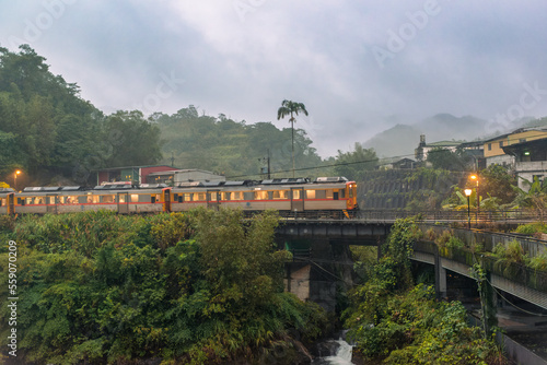 A yellow train runs on the bridge in the evening. A stream in a misty valley. The train station is located in Wanggu, New Taipei City, Taiwan.