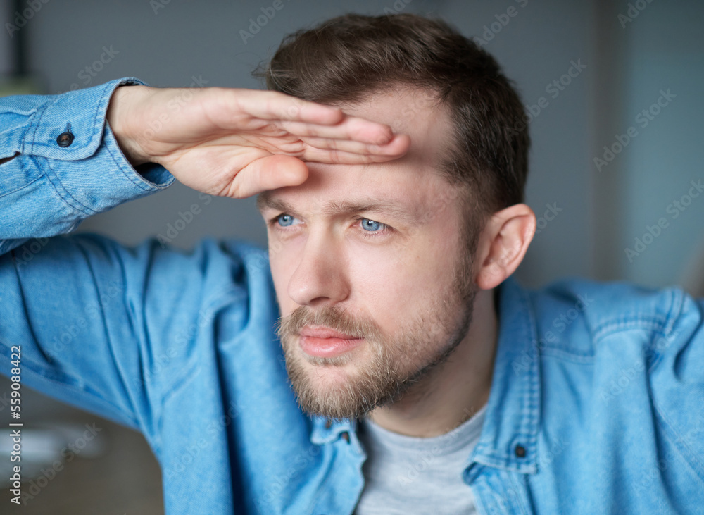 Fototapeta premium Attractive bearded portrait freelance male software engineer at working place in office looking forward for new opportunities with computer on background. Hand near forehead gesture