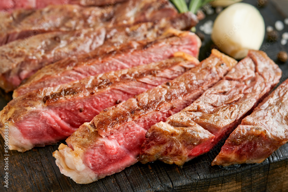 Steaks. Sliced grilled meat steak New York or Ribeye with spices rosemary and pepper on black marble board on old wooden background. Top view. Mock up.