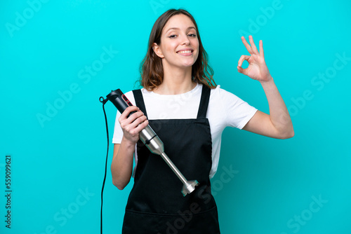 Young caucasian cooker woman using hand blender isolated on blue background showing ok sign with fingers