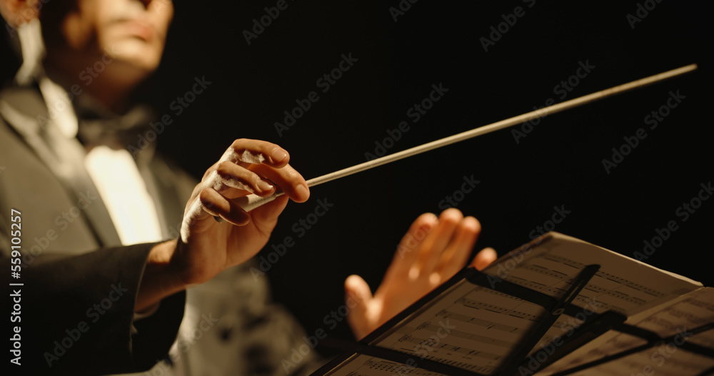 Obraz premium Male orchestra conductor controlling music in orchestra pit by movement of his hands and white baton, studio shot on black background 