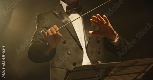 Male orchestra conductor wearing tux standing in front of music stand, controlling musicians by moving his hands and baton. Studio shot on black background  photo