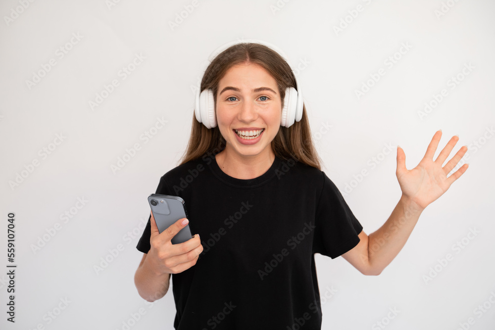 Fototapeta premium Portrait of happy young woman in headphones holding smartphone. Caucasian lady wearing black T-shirt listening to mobile mp3 player over white background. Music and mobile app concept