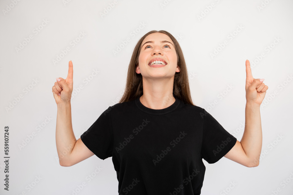 Portrait of happy young woman pointing upwards over white background. Caucasian lady wearing black T-shirt advertising something. Promotion concept Portrait of happy young woman pointing upwards over white background. Caucasian lady wearing black T-shirt advertising something. Promotion concept