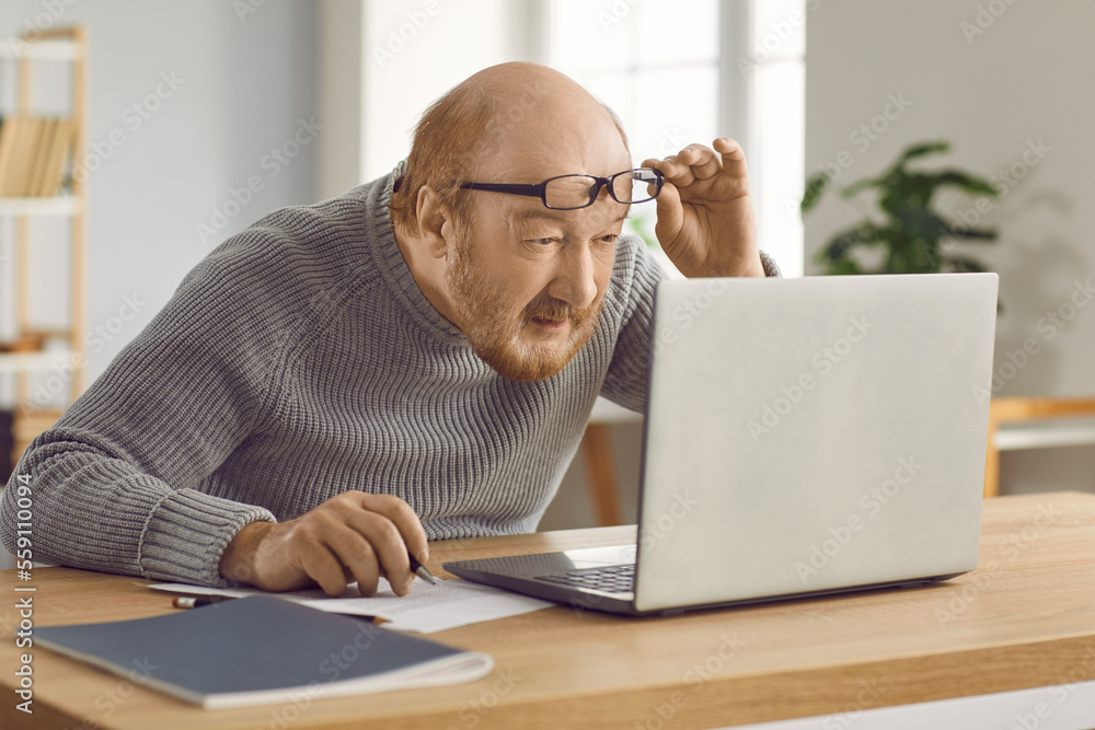 Eyesight problems. Confused mature man who has poor eyesight and works on  laptop at home wearing