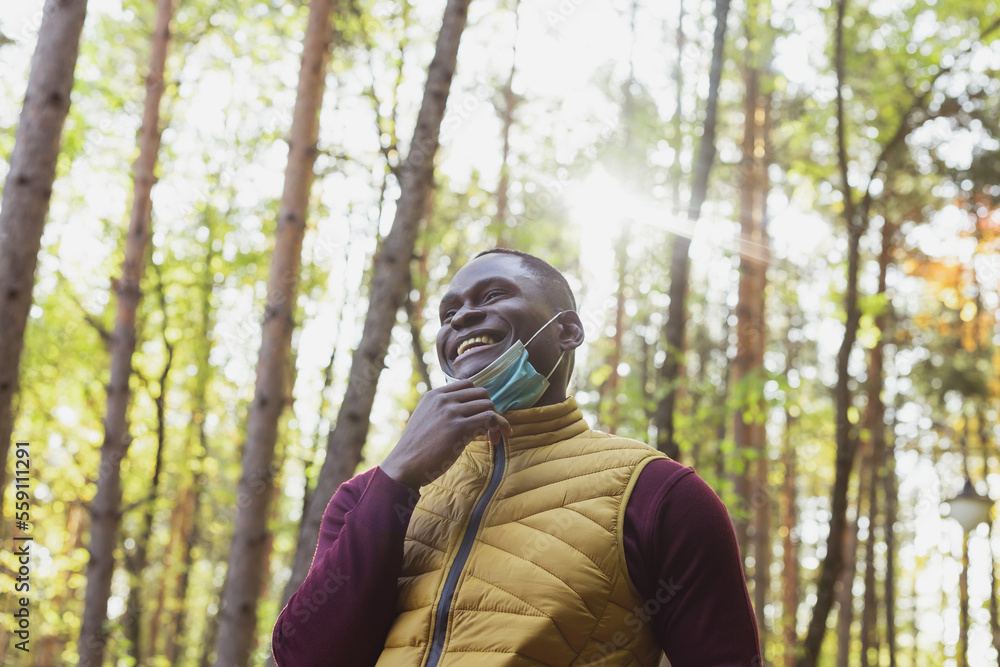 Handsome african american man wearing casual clothes take off medical mask smiling happy walking in city park - end of pandemic Handsome african american man wearing casual clothes take off medical mask smiling happy walking in city park - end of pandemic