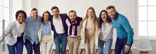 Diverse team of happy united business people hugging each other in the office. Mixed race group of young men and women standing in row  huddling  smiling and laughing together. Unity  teamwork concept