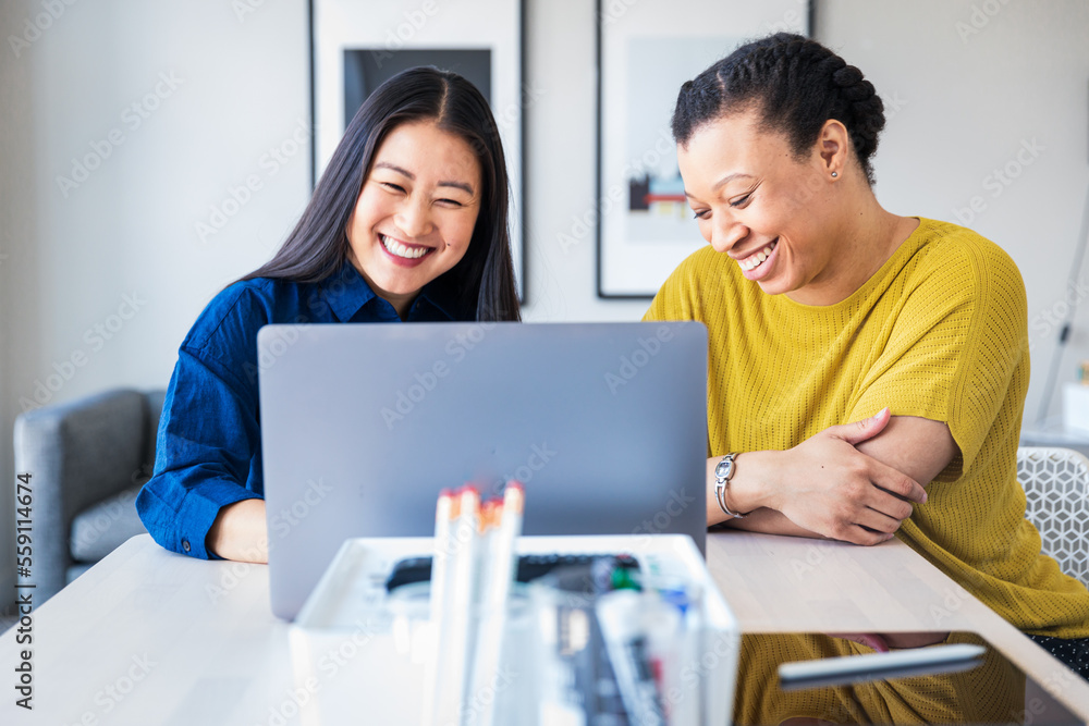 Smiling female coworkers discussing over laptop at table in office Smiling female coworkers discussing over laptop at table in office