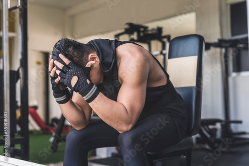 A tanned man sitting on an incline bench, leaning down and covering his head in frustration. Experiencing a workout plateau or lack of gains.