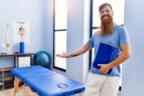 Young redhead man wearing physiotherapist uniform holding clipboard at physiotherapy clinic