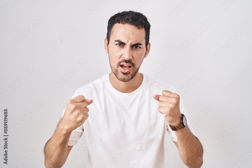 Handsome hispanic man standing over white background angry and mad raising fists frustrated and furious while shouting with anger. rage and aggressive concept. Handsome hispanic man standing over white background angry and mad raising fists frustrated and furious while shouting with anger. rage and aggressive concept.