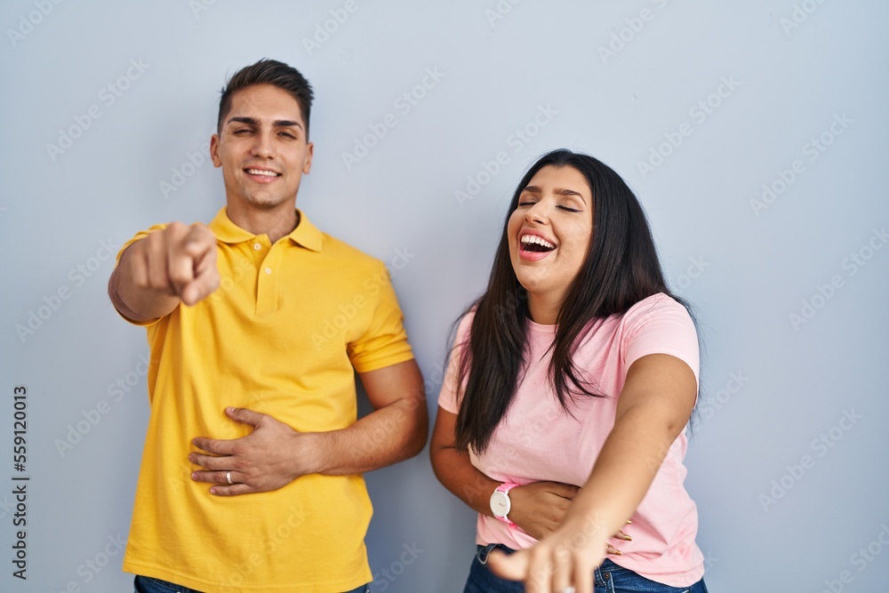 Young couple standing over isolated background laughing at you, pointing finger to the camera with hand over body, shame expression Young couple standing over isolated background laughing at you, pointing finger to the camera with hand over body, shame expression