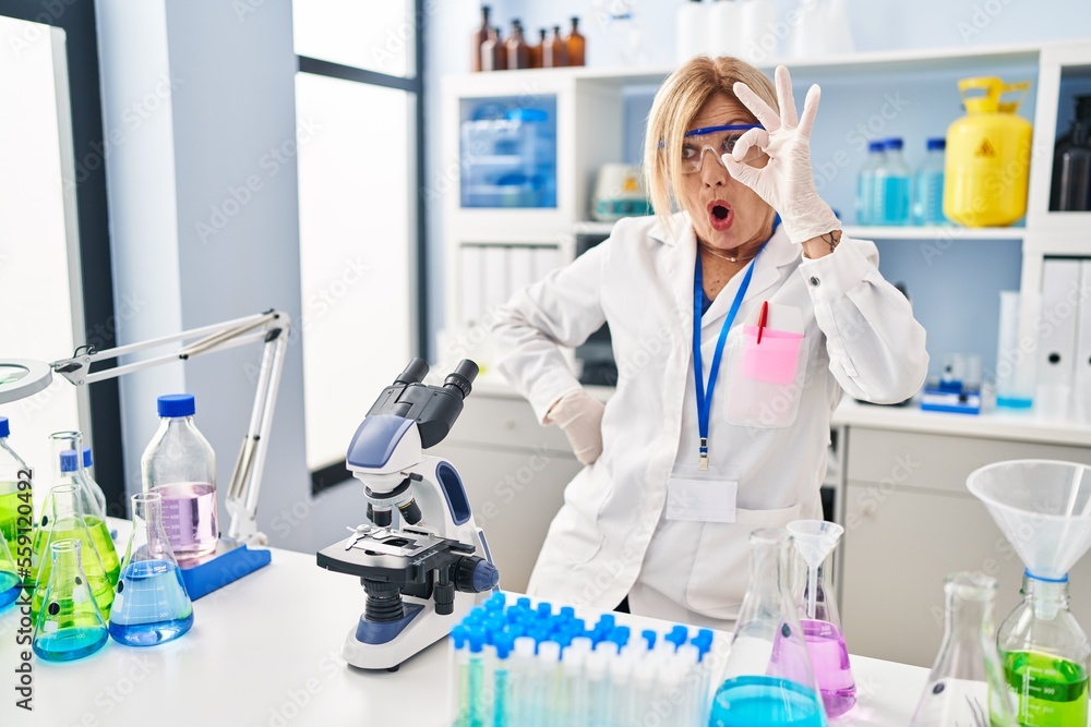 Middle age blonde woman working at scientist laboratory doing ok gesture shocked with surprised face, eye looking through fingers. unbelieving expression. Middle age blonde woman working at scientist laboratory doing ok gesture shocked with surprised face, eye looking through fingers. unbelieving expression.