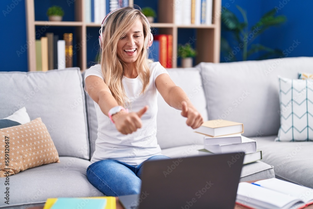 Young blonde woman studying using computer laptop at home approving doing positive gesture with hand, thumbs up smiling and happy for success. winner gesture. Young blonde woman studying using computer laptop at home approving doing positive gesture with hand, thumbs up smiling and happy for success. winner gesture.