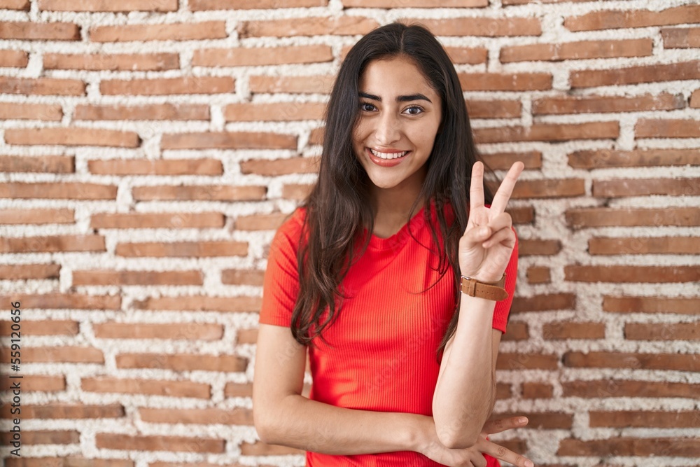 Young teenager girl standing over bricks wall smiling with happy face winking at the camera doing victory sign with fingers. number two. Young teenager girl standing over bricks wall smiling with happy face winking at the camera doing victory sign with fingers. number two.