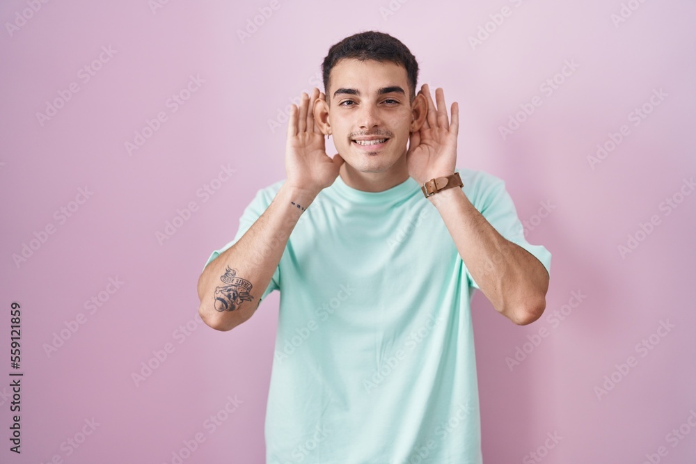 Handsome hispanic man standing over pink background trying to hear both hands on ear gesture, curious for gossip. hearing problem, deaf Handsome hispanic man standing over pink background trying to hear both hands on ear gesture, curious for gossip. hearing problem, deaf