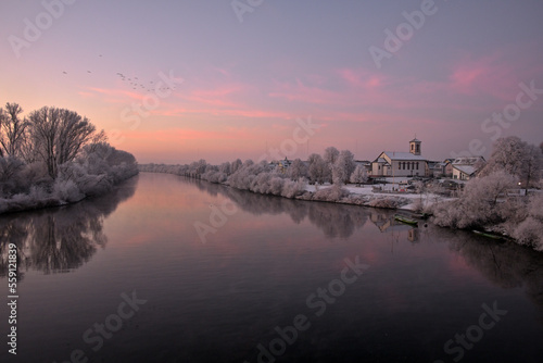 Eine Winterlandschaft am Main zum Morgenrot wo Wildgänse am Himmel fliegen und Bäume mit Raureif und Schnee bedeckt sind. Auf der rechten Bildseite sieht man die Kirche von Mainflingen / Mainhauen.