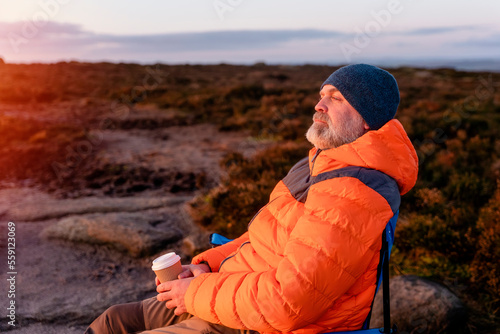 Bearded  Man in orange jacket relaxing alone on the top of  mountain  and drinking hot coffee at sunrise. Travel  Lifestyle concept The national park Peak District in England photo