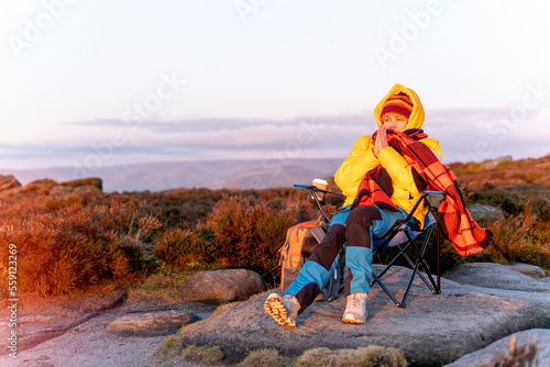 Woman in yellow coat and orange scarf reaching the destination and sitting on beach chairs  on top of hill or mountain at sunrise. British cold winter. Local tourism concept.