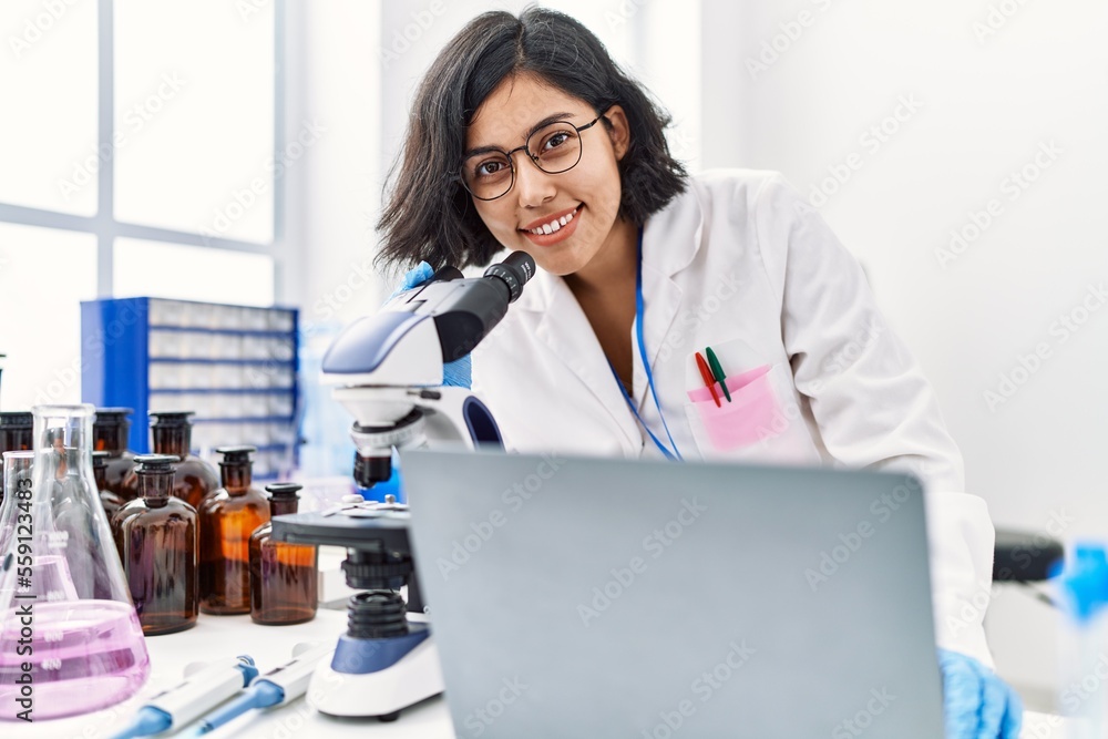 Young latin woman wearing scientist uniform using laptop and microscope at laboratory
