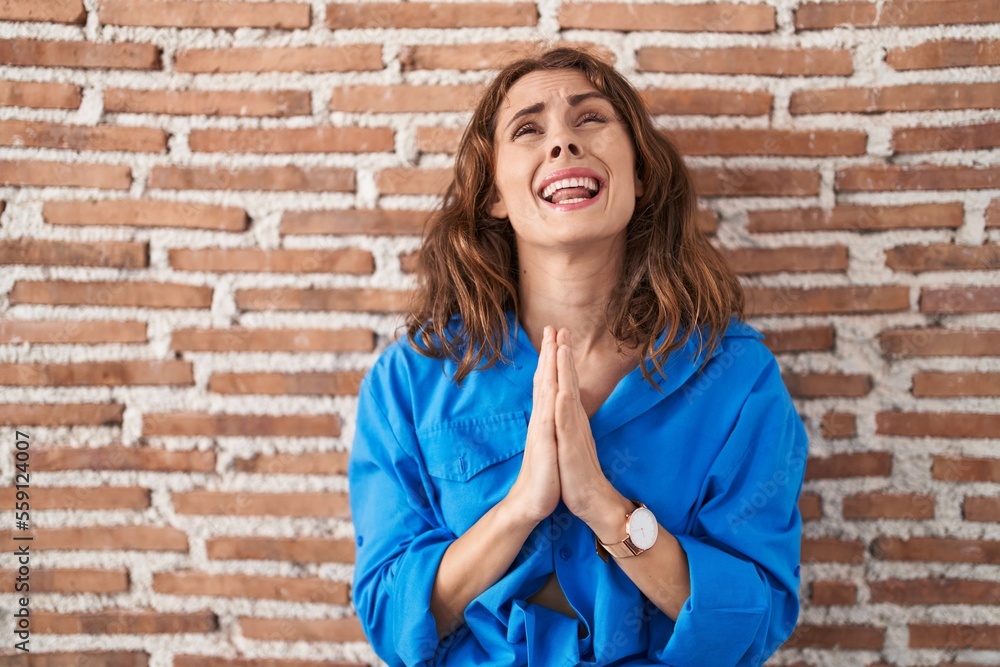 Beautiful brunette woman standing over bricks wall begging and praying with hands together with hope expression on face very emotional and worried. begging. Beautiful brunette woman standing over bricks wall begging and praying with hands together with hope expression on face very emotional and worried. begging.