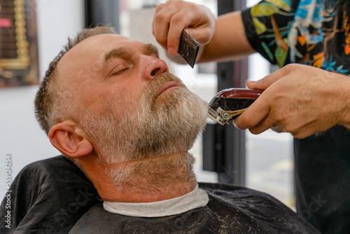 Barber trimming and cutting bearded man with shaving machine in barbershop. Hairstyling process.