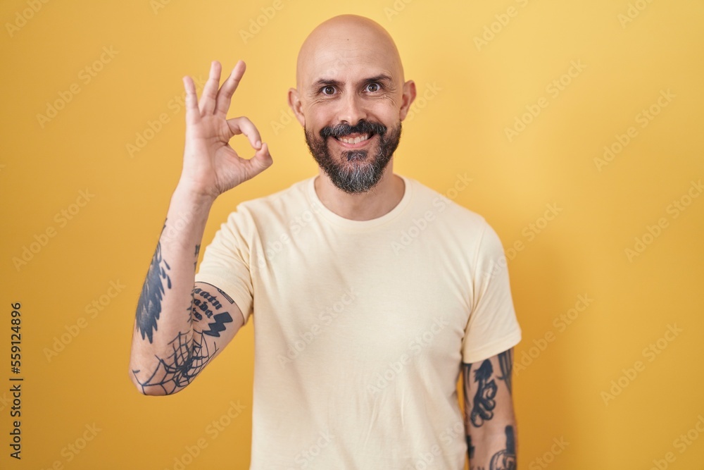 Hispanic man with tattoos standing over yellow background smiling positive doing ok sign with hand and fingers. successful expression. Hispanic man with tattoos standing over yellow background smiling positive doing ok sign with hand and fingers. successful expression.