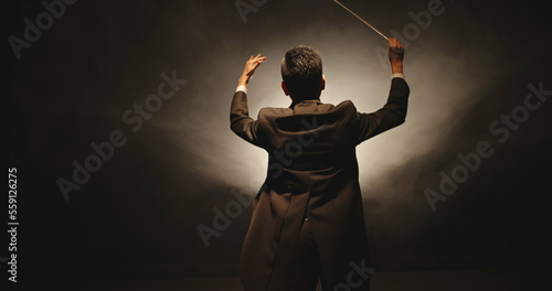 Unrecognizable Male orchestra conductor controlling music in orchestra pit by movement of his hands and white baton, studio shot on black background 