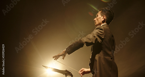 Asian male orchestra conductor wearing tux is directing symphony orchestra with movement of his hands and stick, studio shot on black background  photo