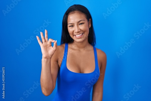 Hispanic woman standing over blue background smiling positive doing ok sign with hand and fingers. successful expression.