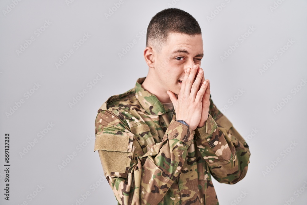Young man wearing camouflage army uniform smelling something stinky and disgusting, intolerable smell, holding breath with fingers on nose. bad smell