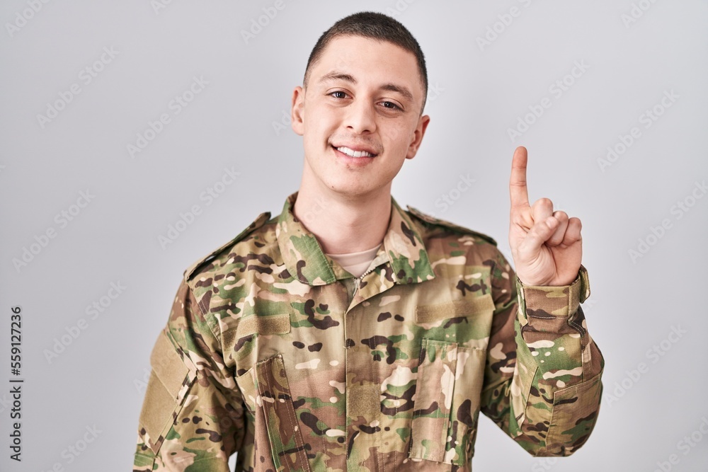 Young man wearing camouflage army uniform showing and pointing up with finger number one while smiling confident and happy. Young man wearing camouflage army uniform showing and pointing up with finger number one while smiling confident and happy.