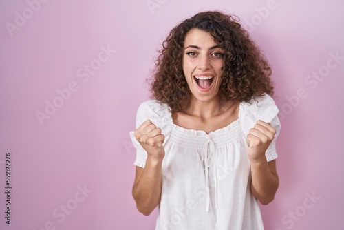 Hispanic woman with curly hair standing over pink background celebrating surprised and amazed for success with arms raised and open eyes. winner concept.