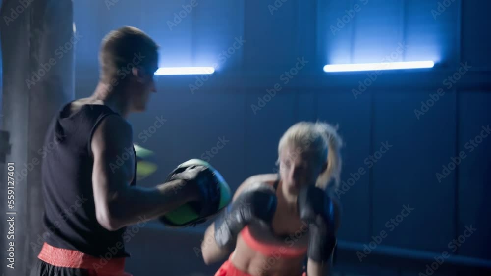 Kickboxing training. Woman in boxing gloves is boxing with her coach in dark gym with blue light. Training sparring of female kickboxer and male trainer. Female fighter practices punches and kicks. Kickboxing training. Woman in boxing gloves is boxing with her coach in dark gym with blue light. Training sparring of female kickboxer and male trainer. Female fighter practices punches and kicks.